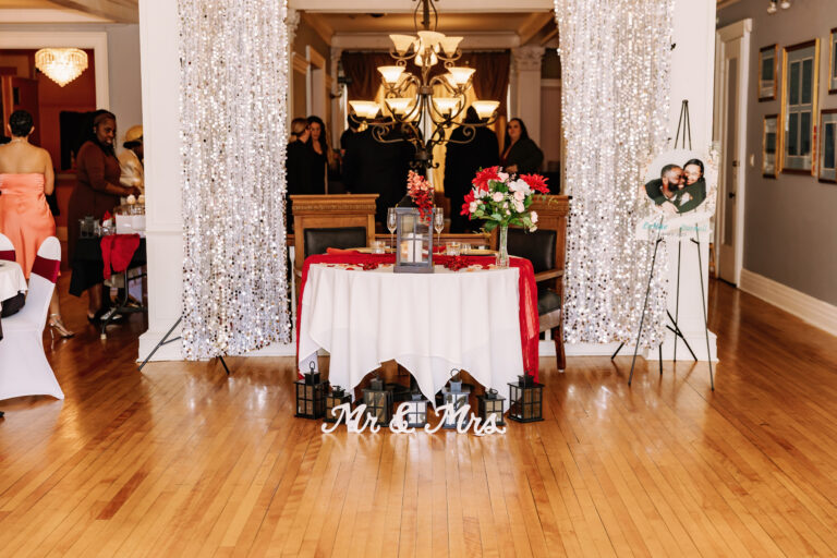 Wedding reception head table with white tablecloth and red runner, floral centerpiece, candles, a "Mr & Mrs" sign on the floor, and guests seated in the background.