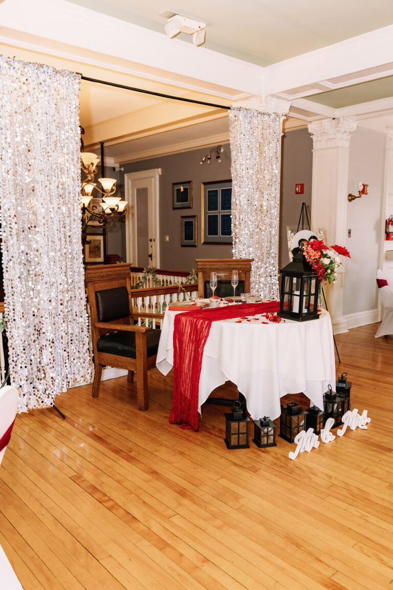 Warmly lit indoor event space with a round table dressed in white cloth and a red runner, lanterns, floral arrangements, and patterned curtains between columns.