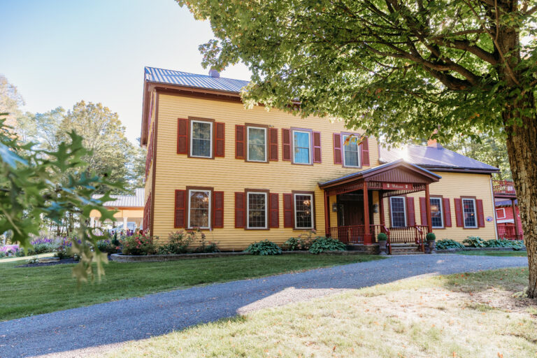 A two-story brick house with red shutters and a small covered front porch sits on a grassy yard with a gravel driveway and surrounding trees.