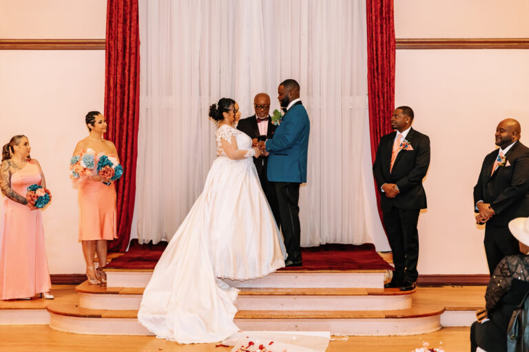 Bride and groom kiss on a wedding stage as bridesmaids in pink dresses and groomsmen in dark suits stand nearby under white drapes and red curtains.