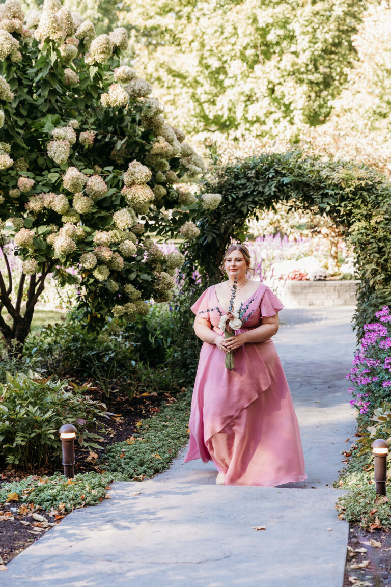 A woman in a pink dress walks along a garden path, holding a bouquet.