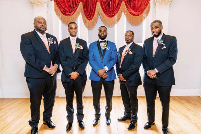 Five men in formal suits posing for a group photo at a wedding, with the center man wearing a blue suit.