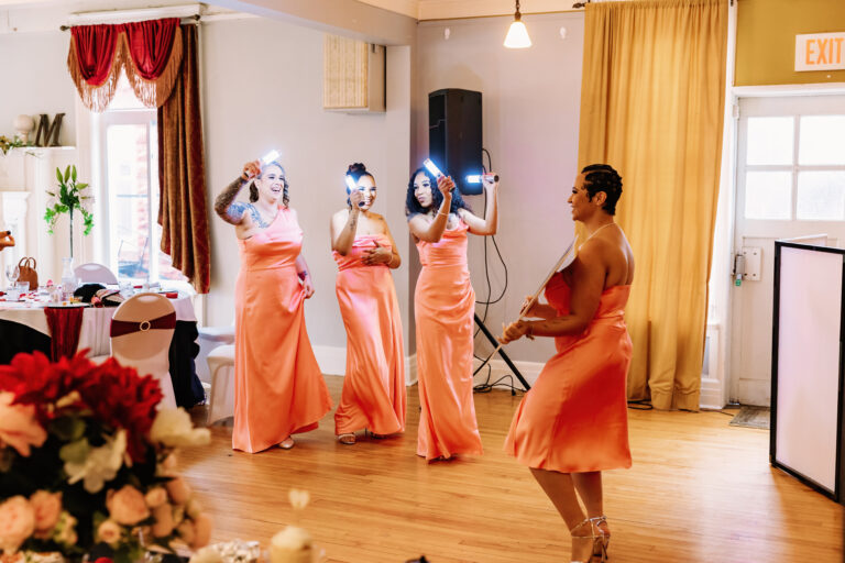 Four women in peach dresses with blue headpieces dance in a decorated room with a wooden floor.
