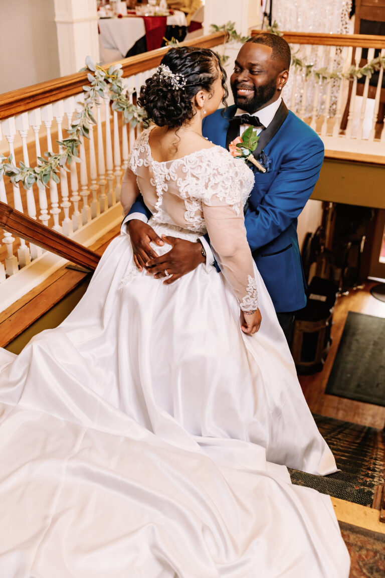 Bride in a white wedding gown with a long train and lace sleeves stands with a groom in a blue suit on a staircase during their wedding.