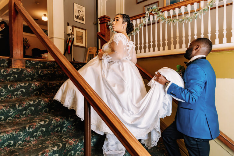 Bride in a white wedding gown on a staircase, assisted by a man in a blue suit.