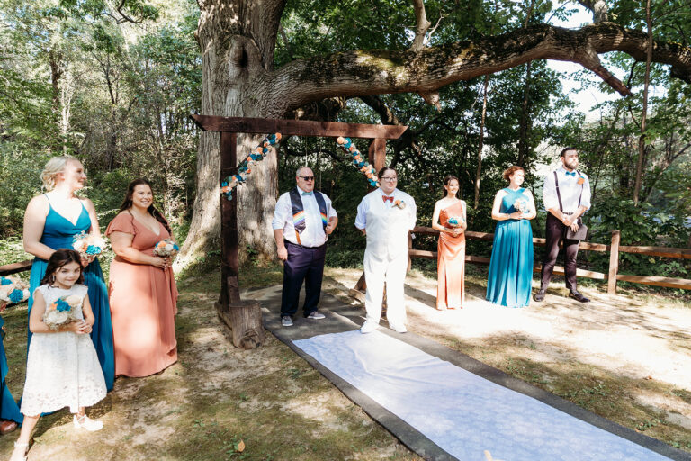Outdoor wedding ceremony under a wooden arch, with the bride and groom flanked by their bridal party along a blue aisle.