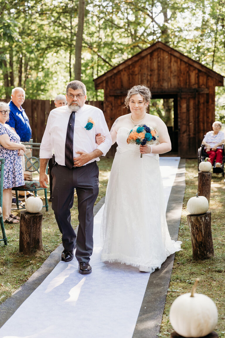 Bride in a white wedding gown walks down an outdoor aisle lined with pumpkins, escorted by a man in a white shirt as guests watch, with a small wooden chapel in the wooded background.