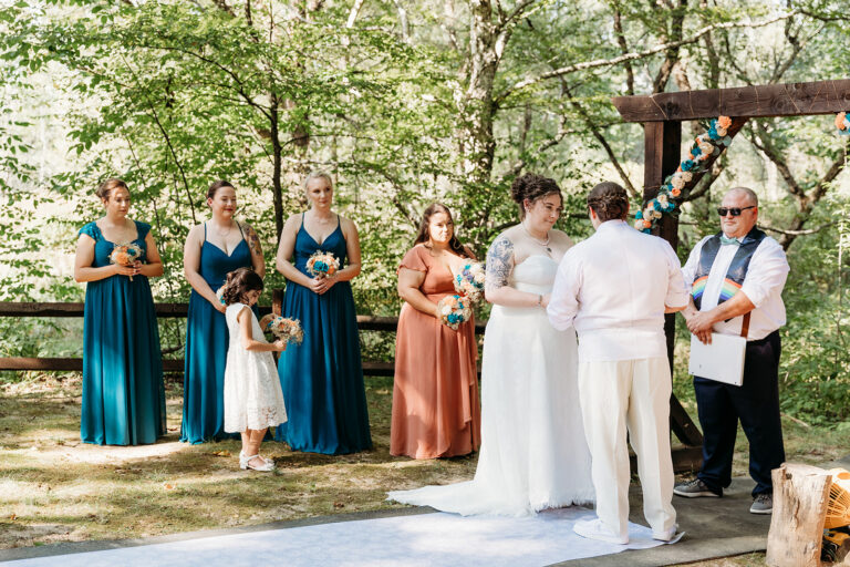 An outdoor wedding ceremony in a wooded setting with the bride and groom at the altar, bridesmaids in teal and rust gowns, a flower girl, and an officiant.