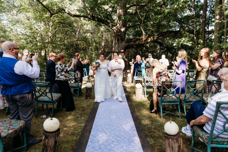 Bride and groom stand at the end of a lavender aisle under trees as guests sit on both sides at an outdoor woodland wedding.