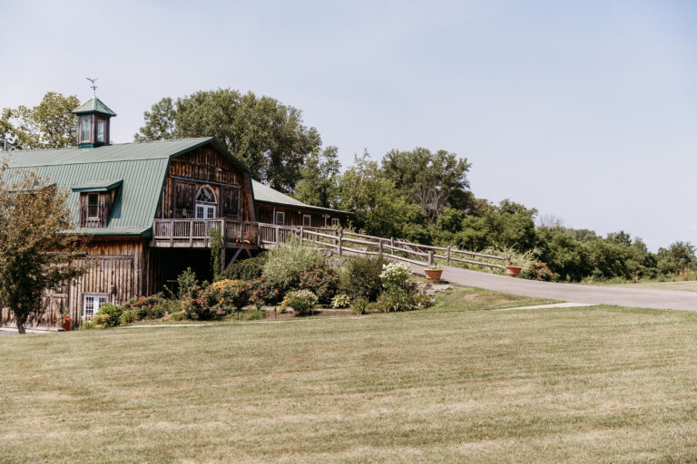 A rustic wooden house with a green roof sits on a grassy hillside, with a winding driveway and trees in the background.