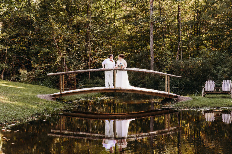 A bride and groom stand on a curved wooden bridge over a calm pond, with their reflection in the water and trees in the background.