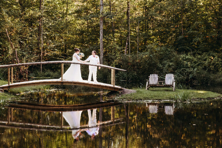 Wedding couple on a curved wooden bridge over a calm pond, their reflection visible in the water among autumn trees.