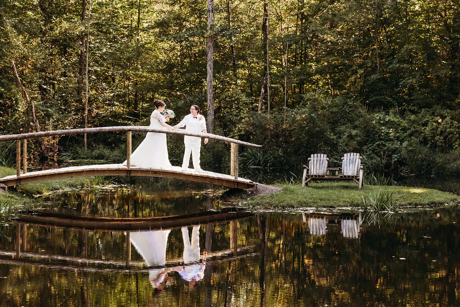 Wedding couple on a curved wooden bridge over a calm pond, their reflection visible in the water among autumn trees.