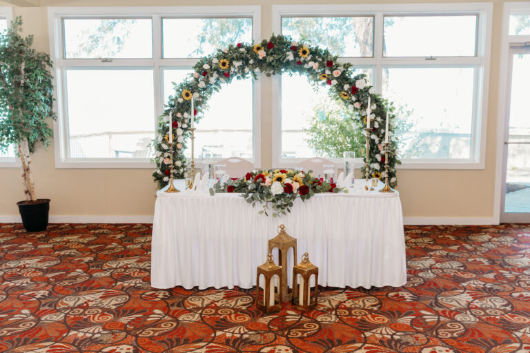 A white-draped table under a circular floral arch with a flower centerpiece and lanterns on the floor in a sunlit room with a red patterned carpet.