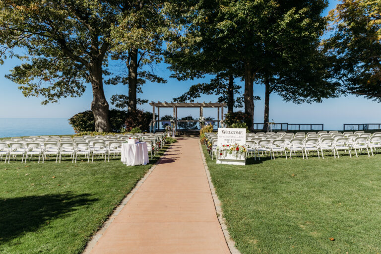 Outdoor lakefront wedding ceremony setup with white folding chairs arranged in rows along a central brick walkway toward a wooden arbor under trees.