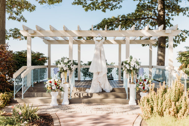Outdoor wedding ceremony site under a white pergola, featuring a wedding gown on display and floral arrangements with a lakeside view.