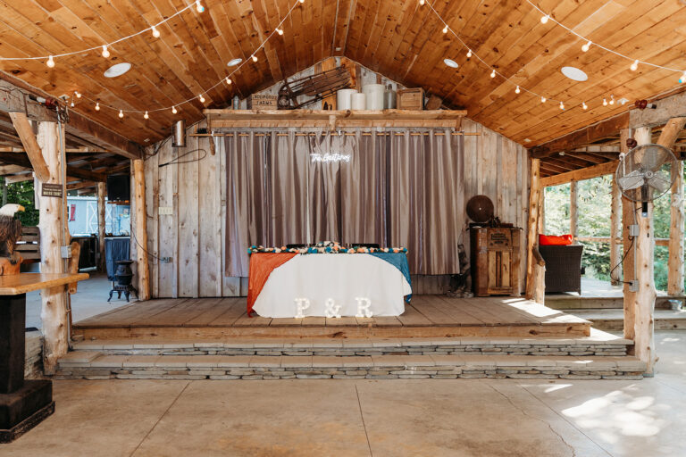 Rustic wooden stage with a long table covered in white cloth, a curtained backdrop, string lights overhead, and a wooden podium on the right.
