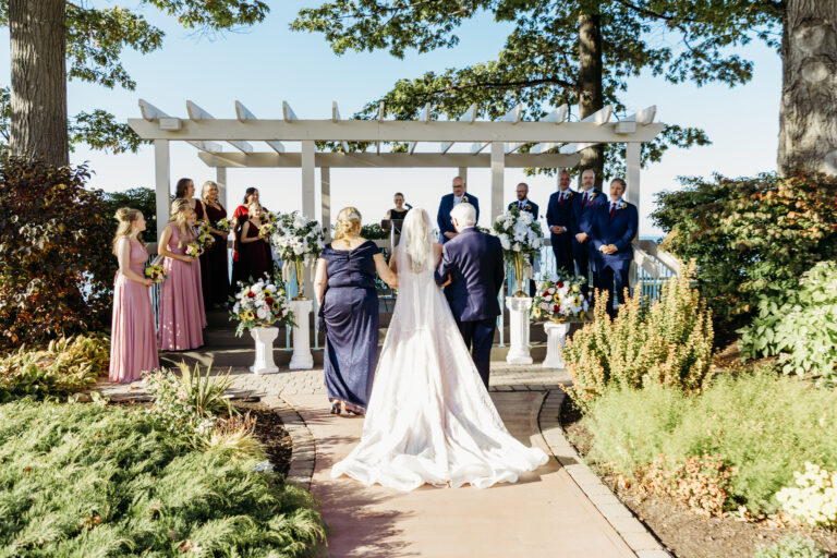 An outdoor garden wedding ceremony with the bride and groom walking toward a white pergola as the wedding party stands nearby.