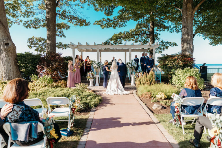 Outdoor wedding ceremony with a couple at a flower-arch altar, guests seated along a flower-lined aisle, under trees with the ocean visible in the background.