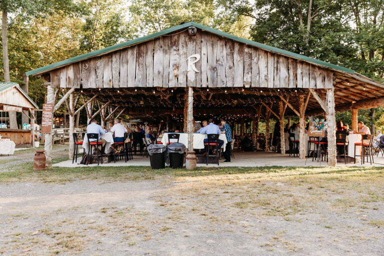 An open-air wooden pavilion with people seated at tables under the shelter in a wooded outdoor area.