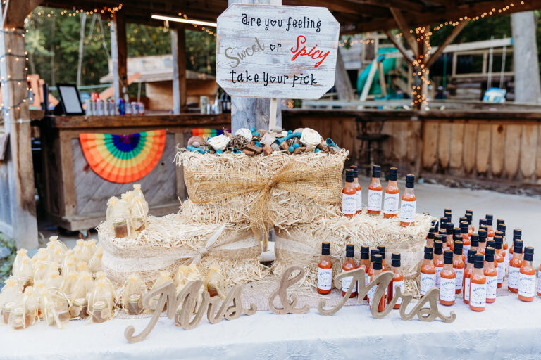Wedding dessert table with a two-tier cake, cupcakes, cake pops, decorative bottles, and a "Mrs & Mrs" sign on a white table under string lights.