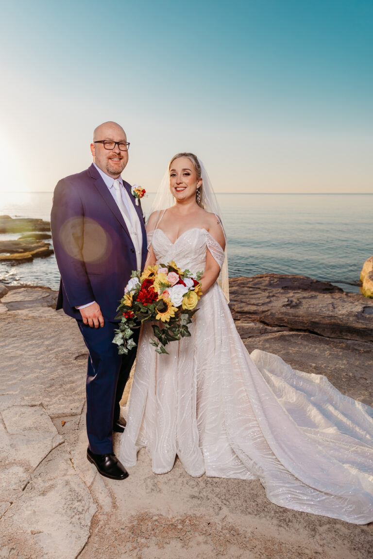 Bride in a white lace wedding gown and veil and groom in a navy suit stand on a rocky shoreline with the sea and sky behind them, the bride holding a colorful bouquet.