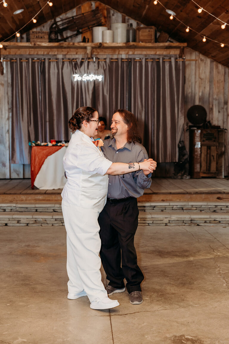 Two people dancing closely in a rustic venue with string lights and a stage in the background.