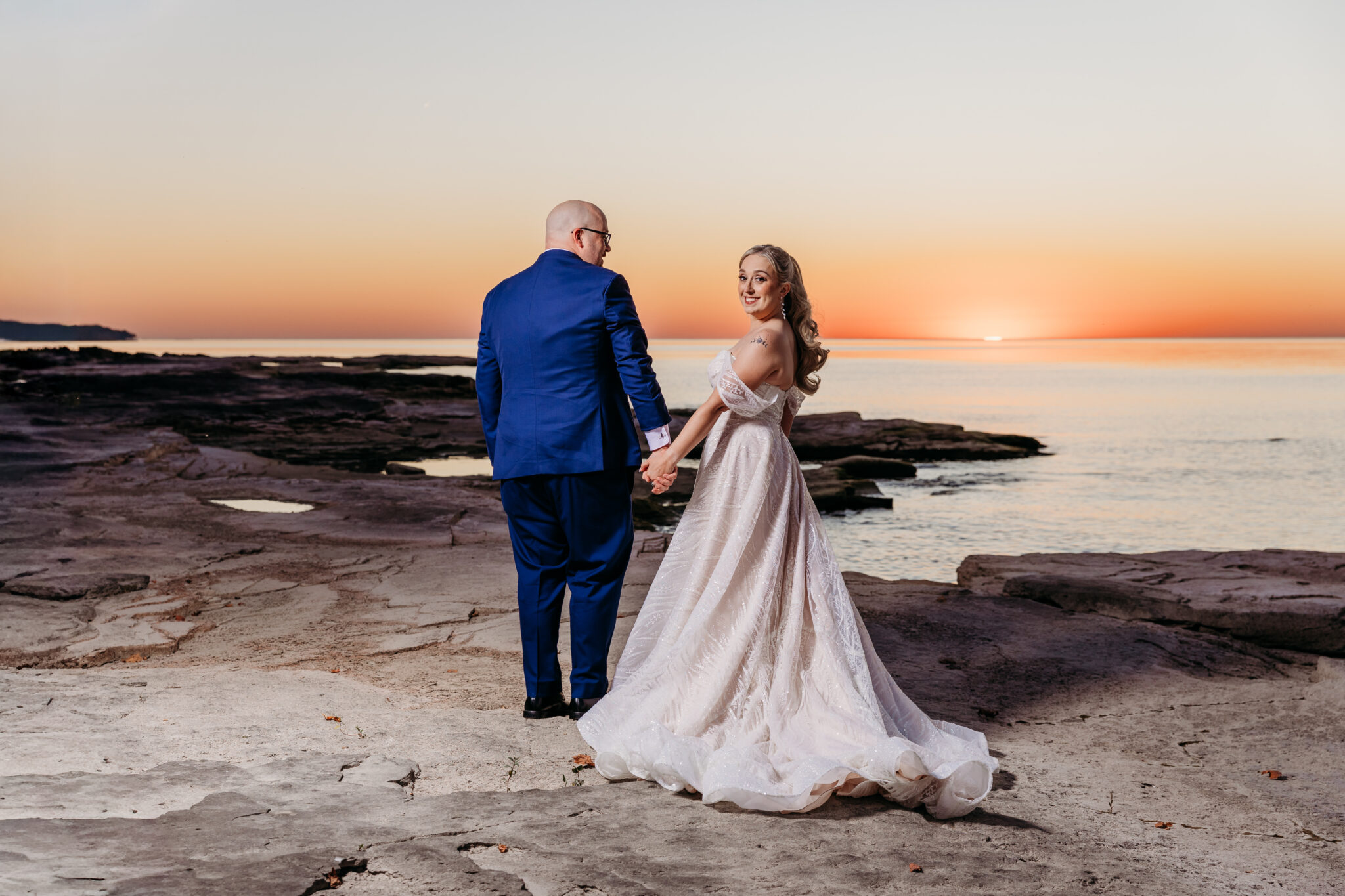 Bride in a white wedding gown and groom in a blue suit stand on a rocky shoreline at sunset, holding hands and looking toward the horizon.
