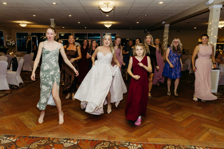 Bride in a white wedding gown leads her bridesmaids in colorful dresses across a ballroom dance floor.
