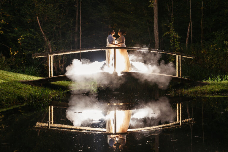Bride and groom embrace on an arched bridge over a calm pond, their reflection visible in the water with wisps of mist around them.