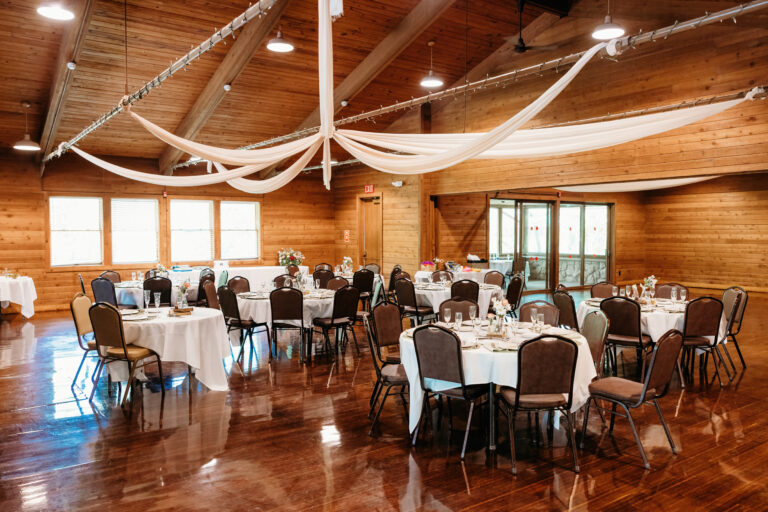 A wooden banquet hall set for a reception with round tables, chairs, white tablecloths, and flowing fabric drapery hanging from the ceiling.