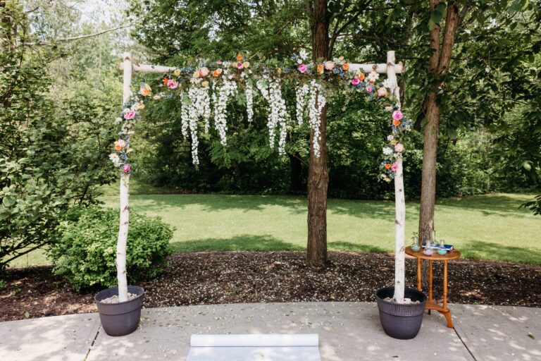 A white floral arch adorned with hanging white blossoms and colorful blooms stands on a stone patio in a garden, flanked by two potted plants.