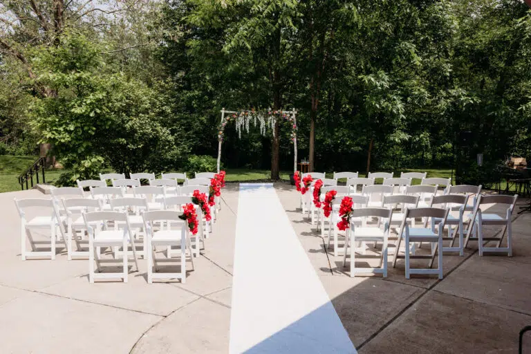 Outdoor wedding ceremony setup with white chairs arranged along a central white aisle and a wooden arch adorned with greenery and red flowers.