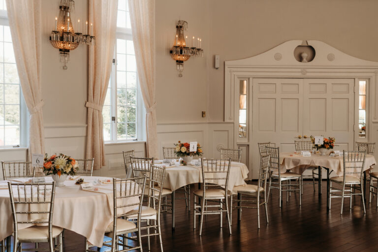 Elegant dining room with round tables covered in white linens, floral centerpieces, gold chairs, and chandeliers, illuminated by large windows in the background.