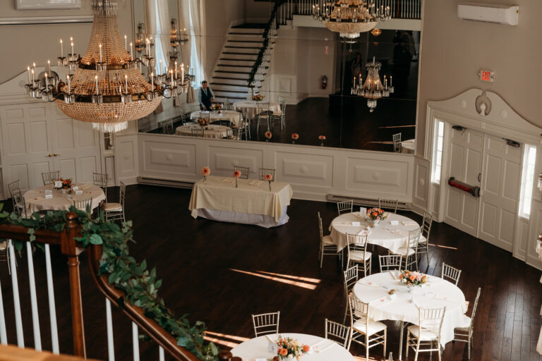 Elegant banquet hall with chandeliers, round tables set with white linens, and a long central table viewed from a staircase landing.