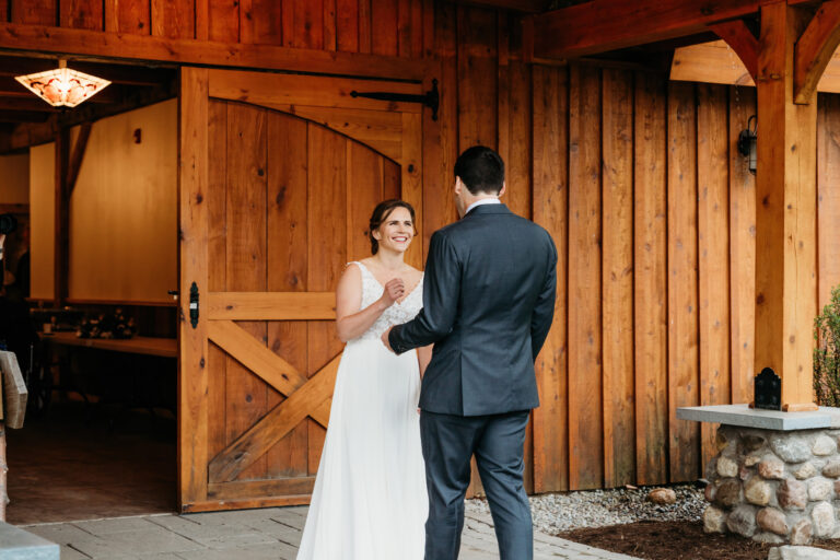 A bride in a white wedding dress and a groom in a dark suit stand facing each other outside a rustic wooden barn, smiling.