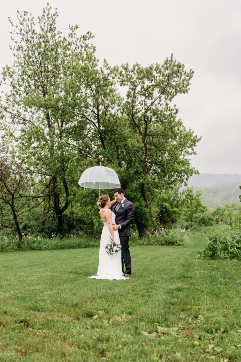 Bride and groom embrace under a white umbrella on a grassy lawn with trees in the background.