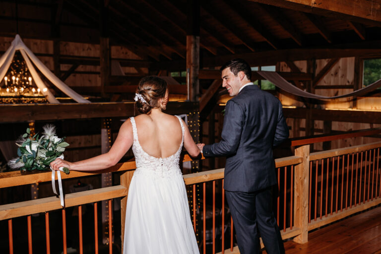 Bride in a white backless wedding dress and her groom in a dark suit stand at a wooden indoor balcony railing, seen from behind.