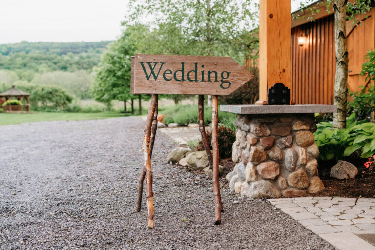 Rustic wooden sign reading "Wedding" on two branch supports beside a gravel path at an outdoor venue.
