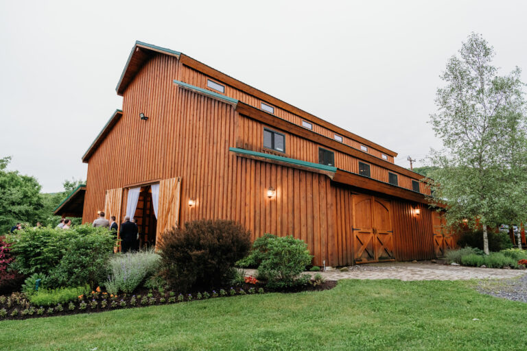 A long brown wooden building with a sloped roof sits among landscaped shrubs and a lawn.