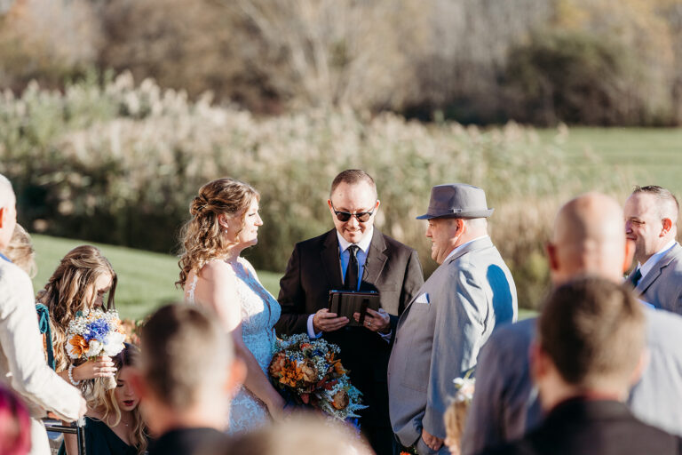 Outdoor wedding ceremony in a sunlit garden with a couple at the center and guests gathered around.
