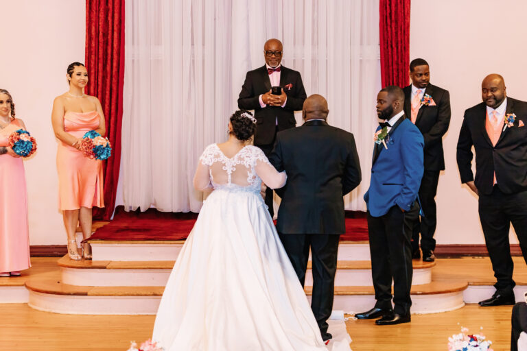 Bride and groom stand before an officiant during a wedding ceremony on a stage, surrounded by bridesmaids in pink dresses and groomsmen in suits.