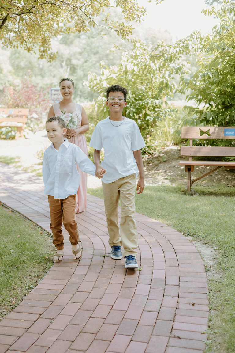 Two children walk along a brick path in a sunny park, with two adults following nearby.