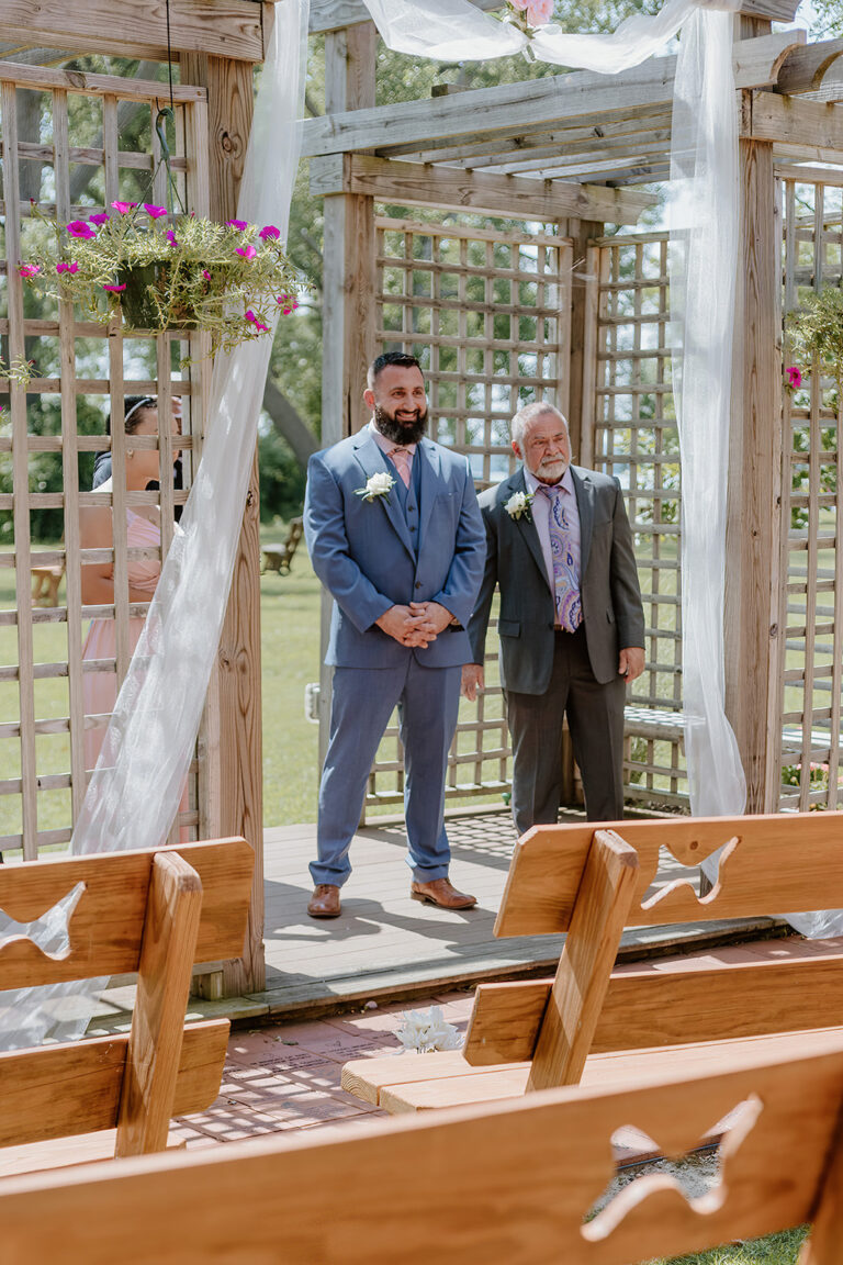 Two men in suits stand under a white-draped wooden arch at an outdoor wedding, with wooden pews and hanging pink flowers visible in the foreground.