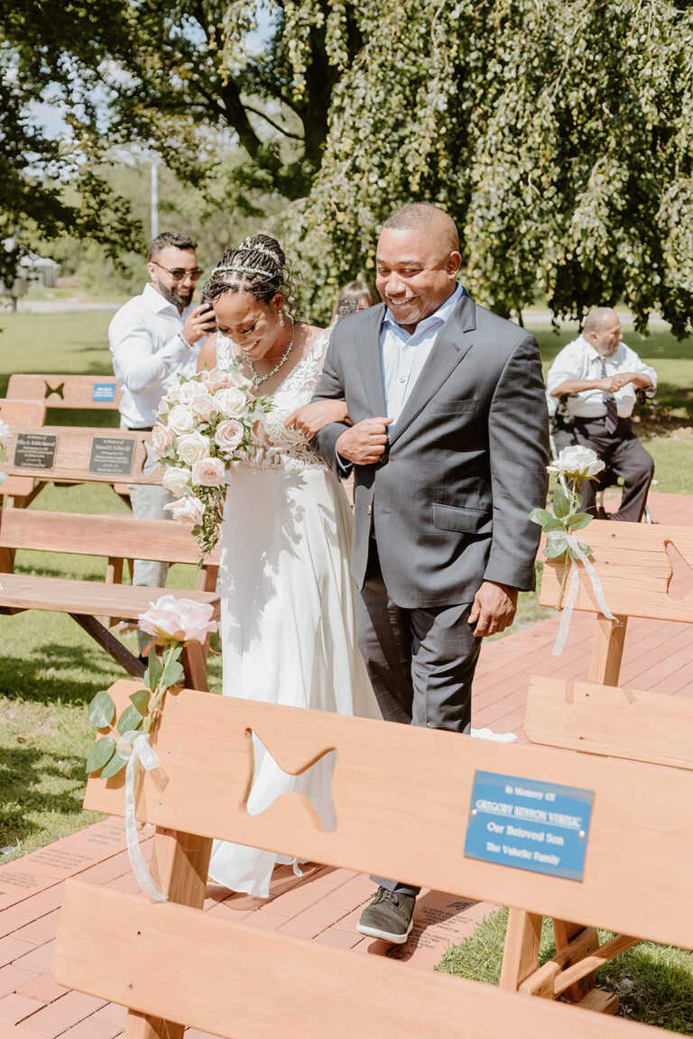 An outdoor wedding where a bride in a white dress holds a bouquet and walks arm-in-arm with a man in a gray suit down a wooden aisle, with guests and trees in the background.