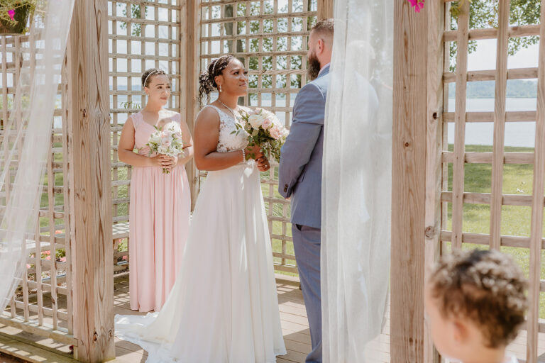 Bride and groom stand under a white-draped wooden gazebo at an outdoor wedding, with a bridesmaid in pink nearby.