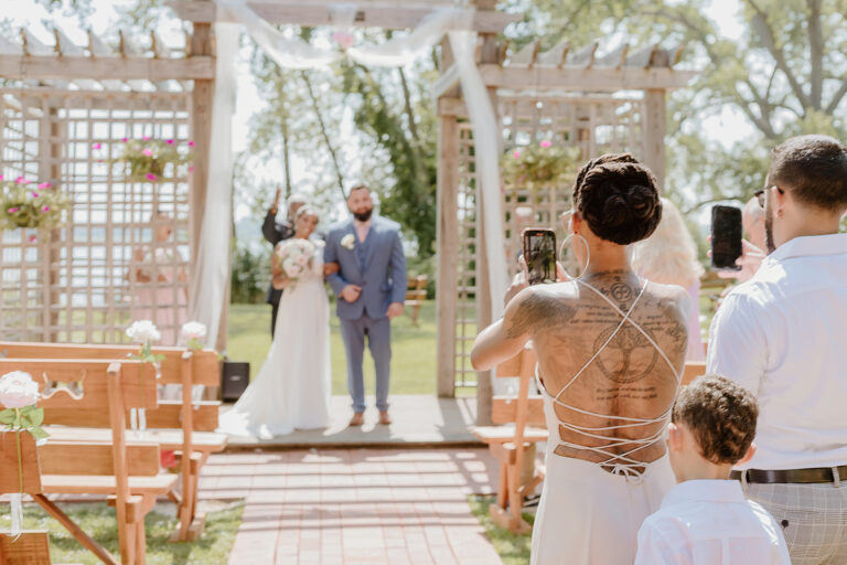 An outdoor wedding ceremony with a bride and groom standing under a wooden arch as guests photograph the moment.
