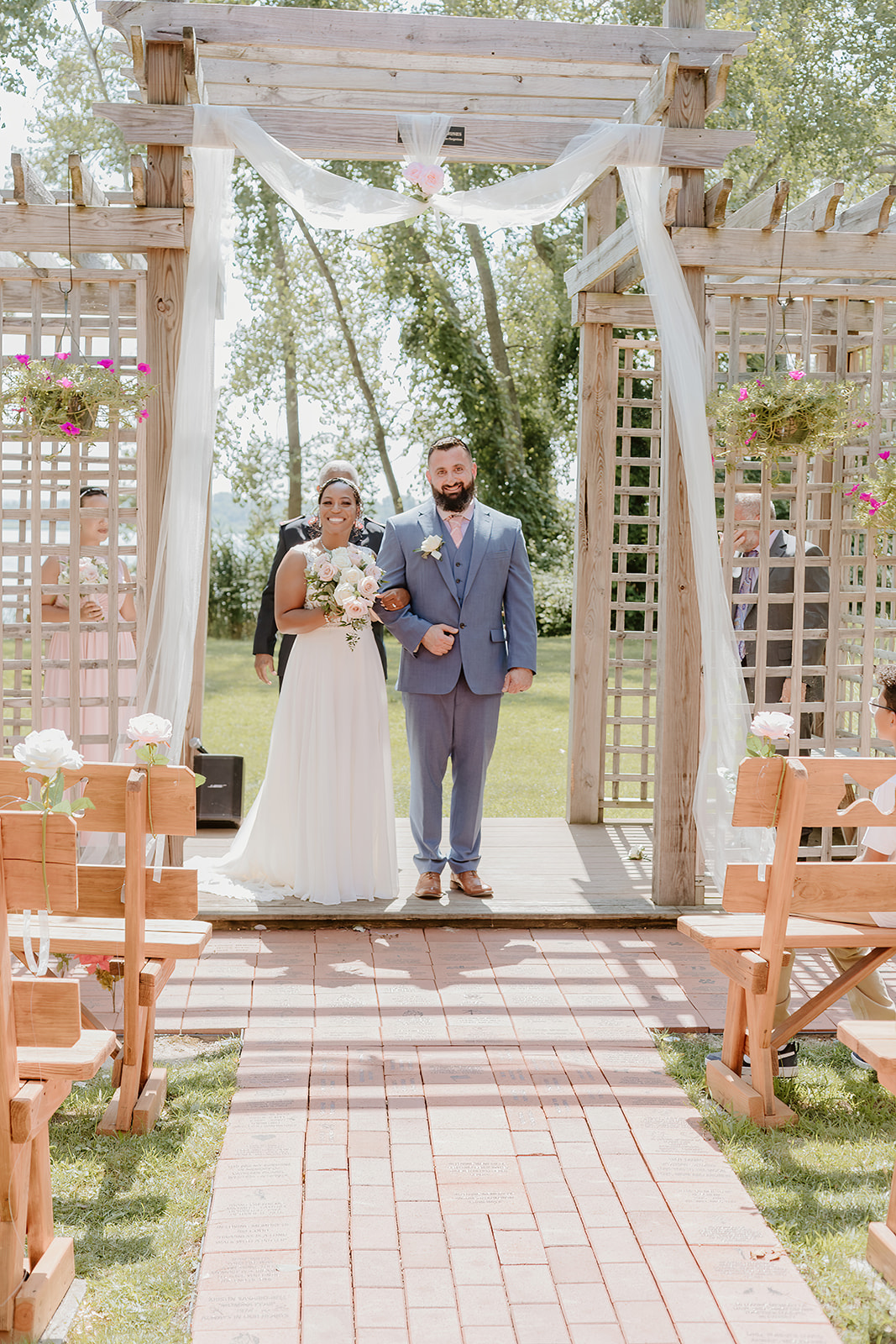 Bride in a white dress and groom in a blue suit walk down a brick-pathed aisle beneath a wooden arch at an outdoor wedding.