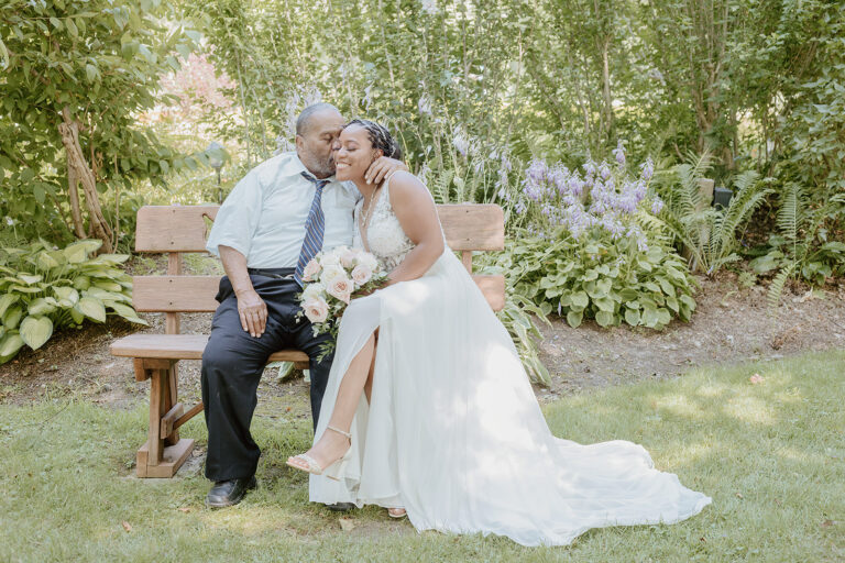 Bride and groom kiss on a wooden bench in a lush garden, with the bride wearing a white wedding dress.