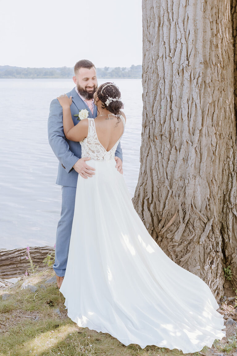 Bride and groom embracing beside a large tree by a lake, the bride in a flowing white gown.